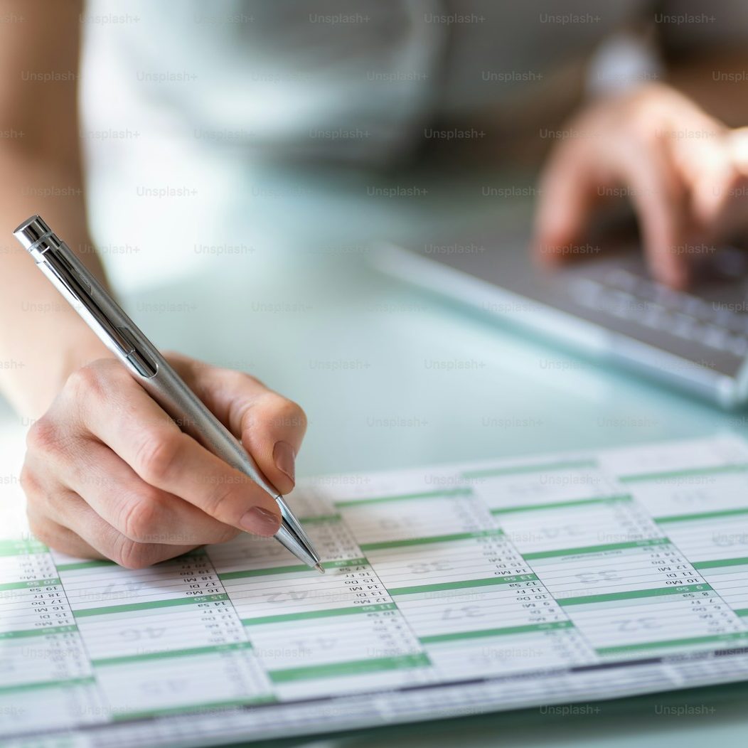 Close-up of a person’s hand holding a pen, writing on a printed calendar or timetable, with the other hand on a laptop keyboard. The image is circular and suggests organisation, planning, or scheduling.